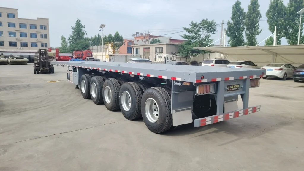 Grey five-axle flatbed semi trailer viewed from the rear corner in a factory yard